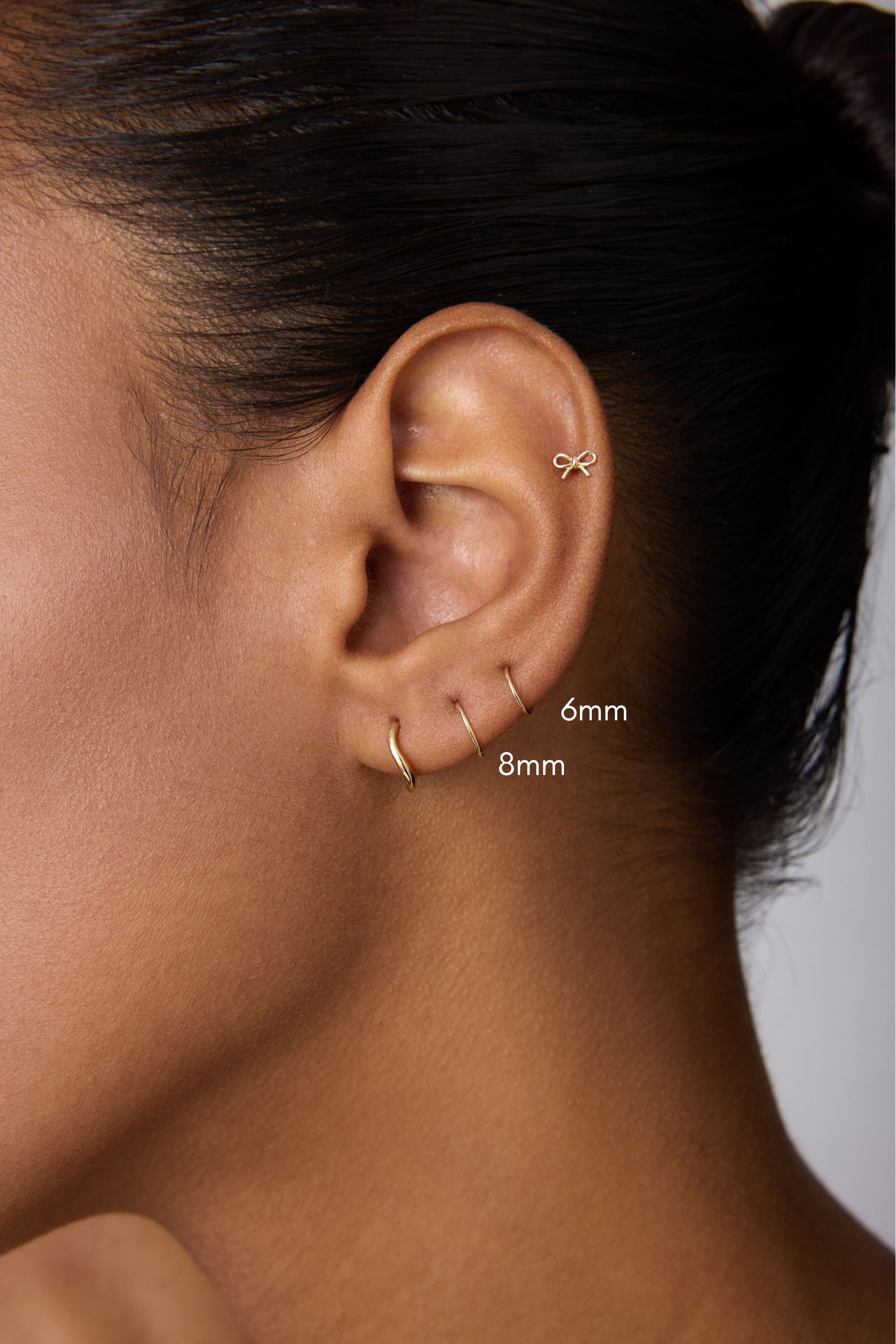 A close-up shows a person’s ear with two 14K Solid Yellow Gold 20G Mini Clickers (6mm and 8mm) in the lobe and a small bow-shaped stud in the upper cartilage, against a neutral background.