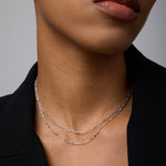 A close-up of a person wearing a black blazer and two delicate silver necklaces, including the Mini Paperclip Double Chain Necklace in Silver, with natural makeup and neutral lips. The image highlights the neck and lower face.
