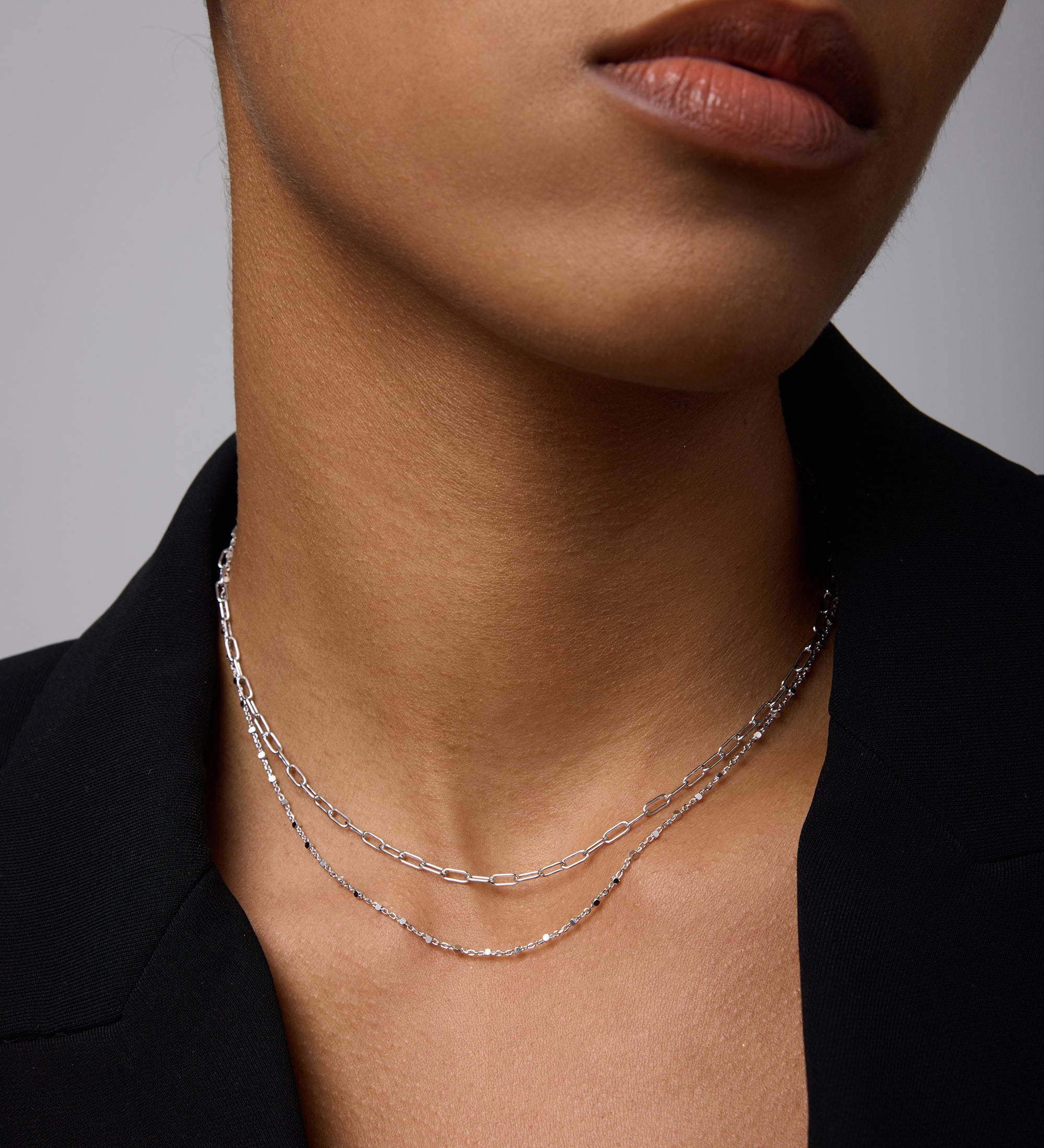 A close-up of a person wearing a black blazer and two delicate silver necklaces, including the Mini Paperclip Double Chain Necklace in Silver, with natural makeup and neutral lips. The image highlights the neck and lower face.