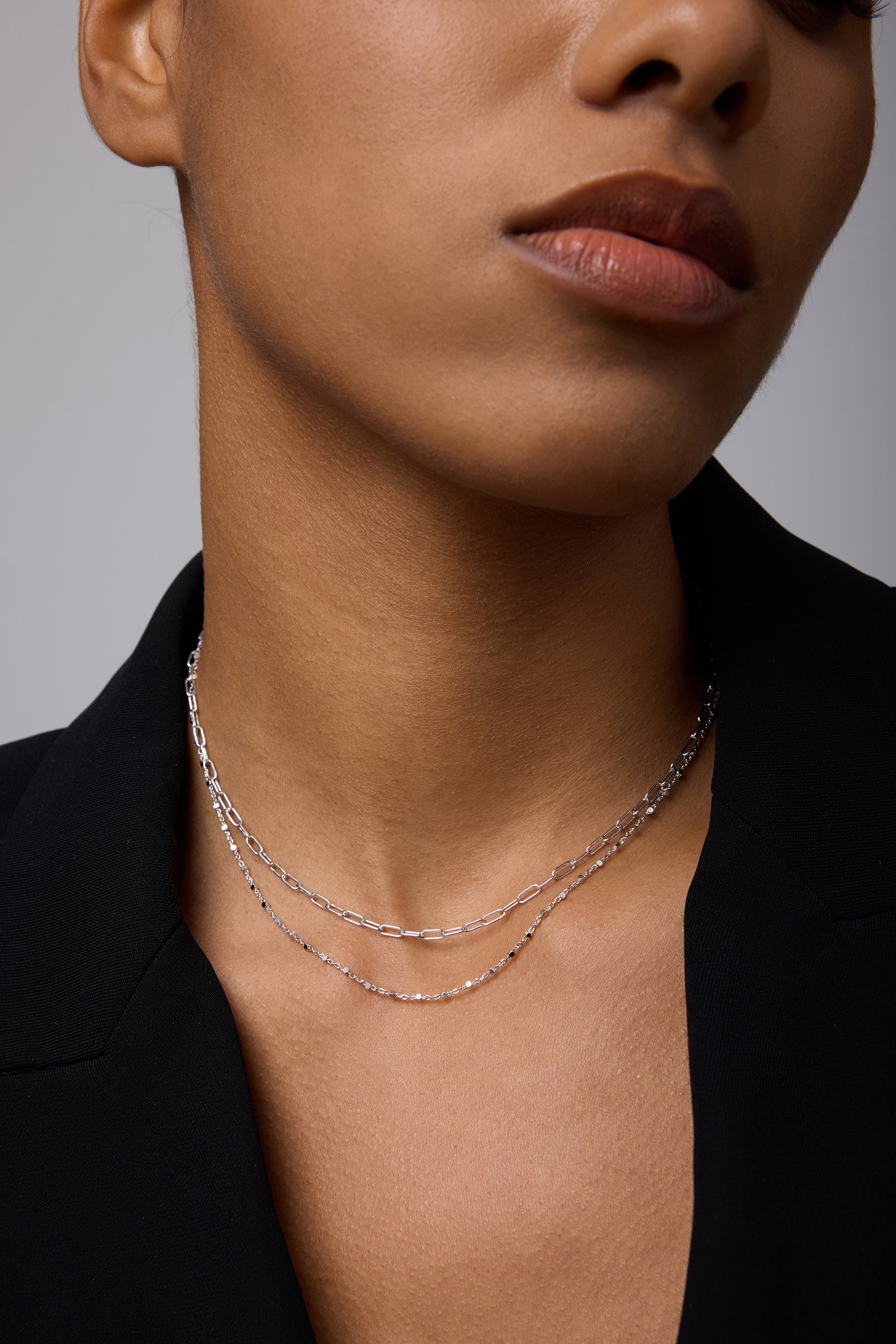 A close-up of a person wearing a black blazer and two delicate silver necklaces, including the Mini Paperclip Double Chain Necklace in Silver, with natural makeup and neutral lips. The image highlights the neck and lower face.