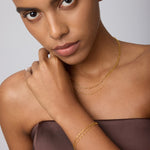 A woman in a strapless brown top touches her neck and looks at the camera, wearing a Mini Paperclip Double Chain Bracelet in Gold with a matching necklace against a light background.