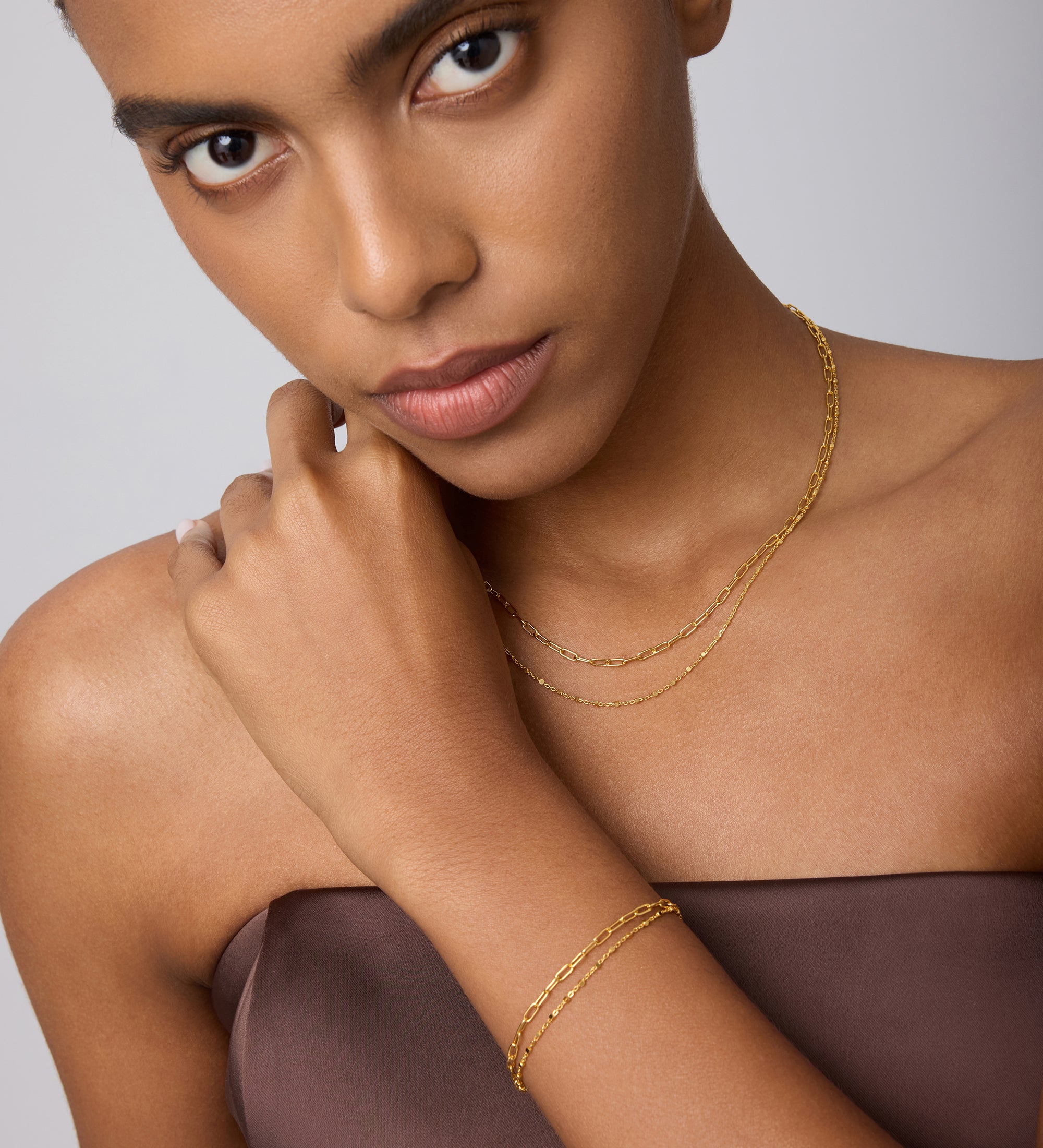 A woman in a strapless brown top touches her neck and looks at the camera, wearing a Mini Paperclip Double Chain Bracelet in Gold with a matching necklace against a light background.