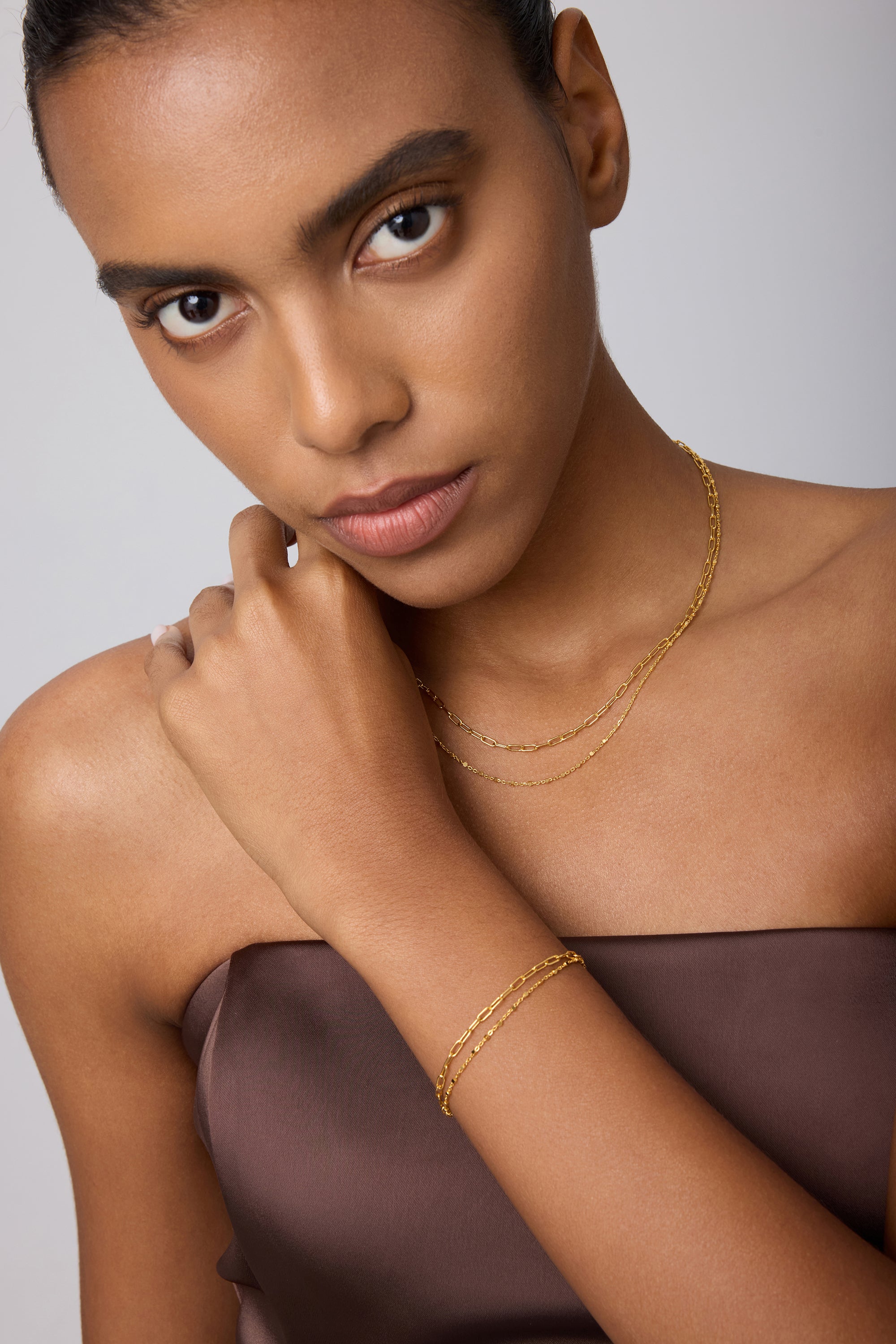 A woman in a strapless brown top touches her neck and looks at the camera, wearing a Mini Paperclip Double Chain Bracelet in Gold with a matching necklace against a light background.