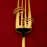 The Molten Crystal Double Band Ring in Gold, featuring two 18k gold plated bands, is elegantly displayed on a golden fork against a deep red background.