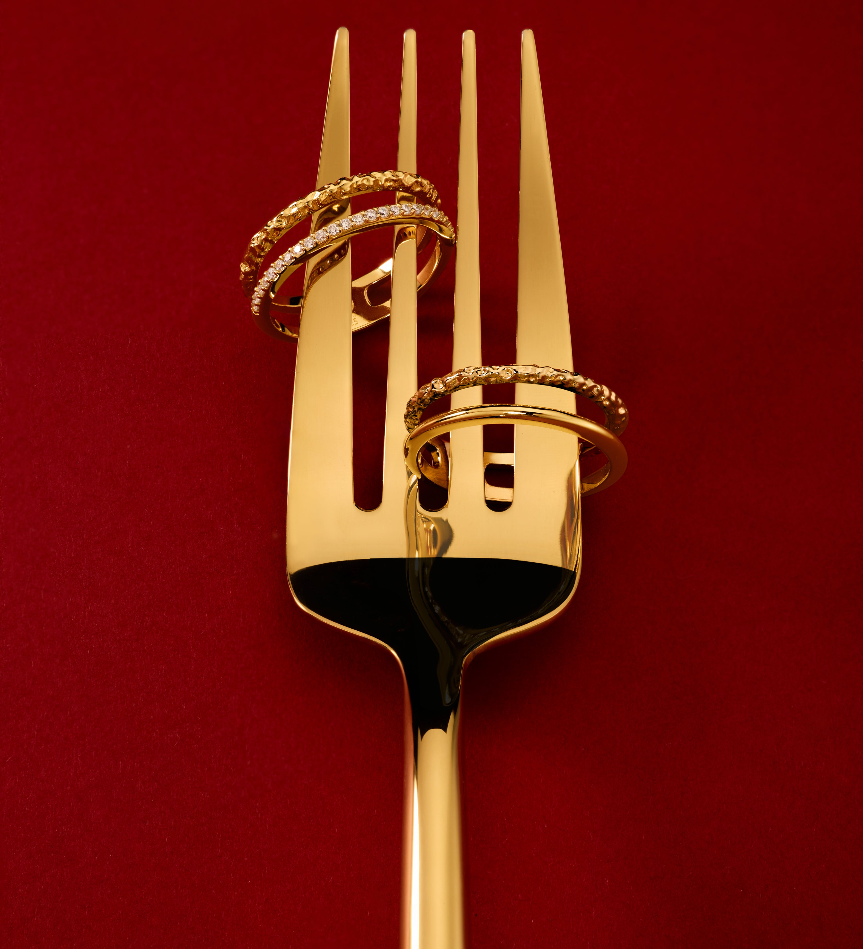 The Molten Crystal Double Band Ring in Gold, featuring two 18k gold plated bands, is elegantly displayed on a golden fork against a deep red background.