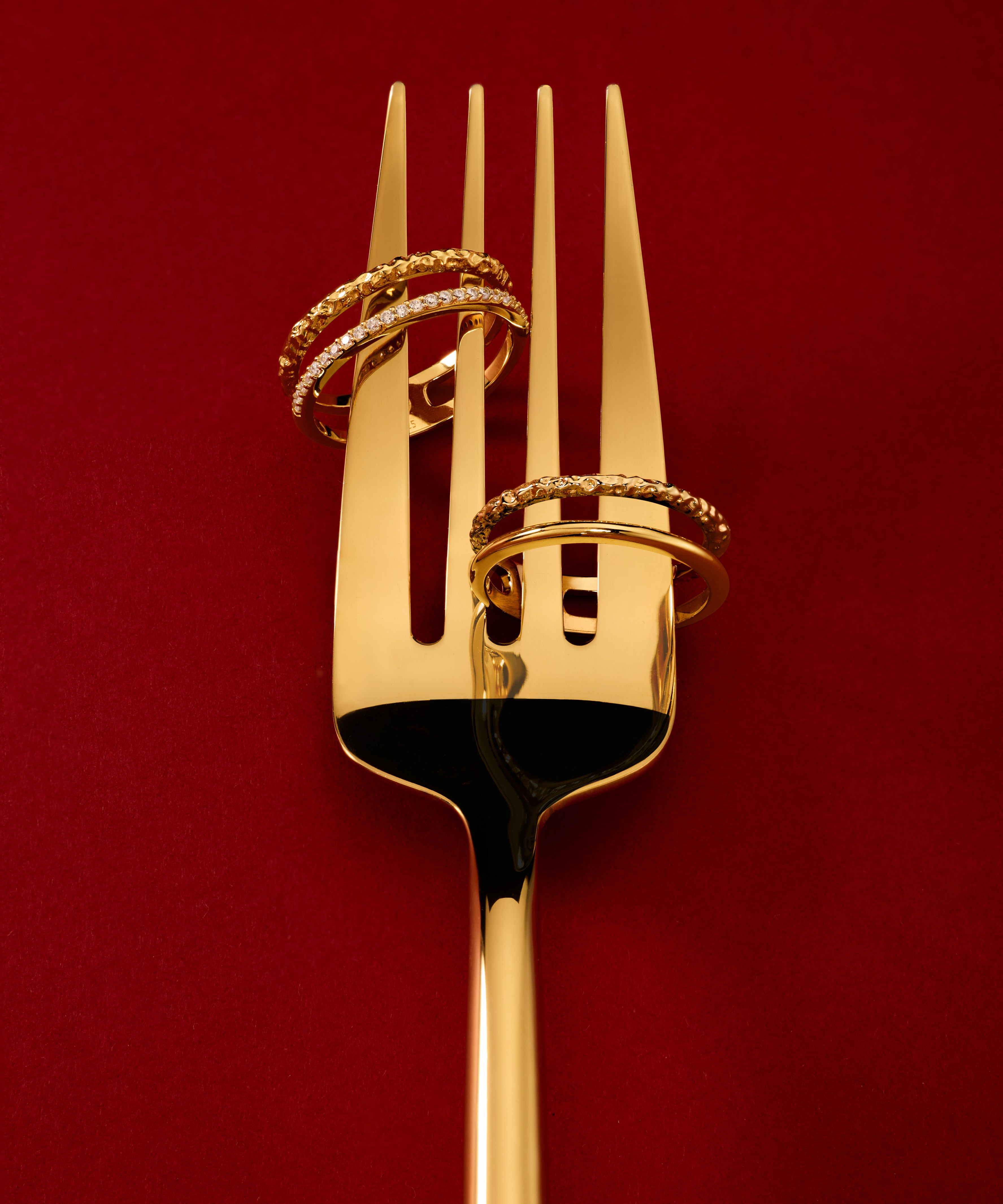 The Molten Crystal Double Band Ring in Gold, featuring two 18k gold plated bands, is elegantly displayed on a golden fork against a deep red background.