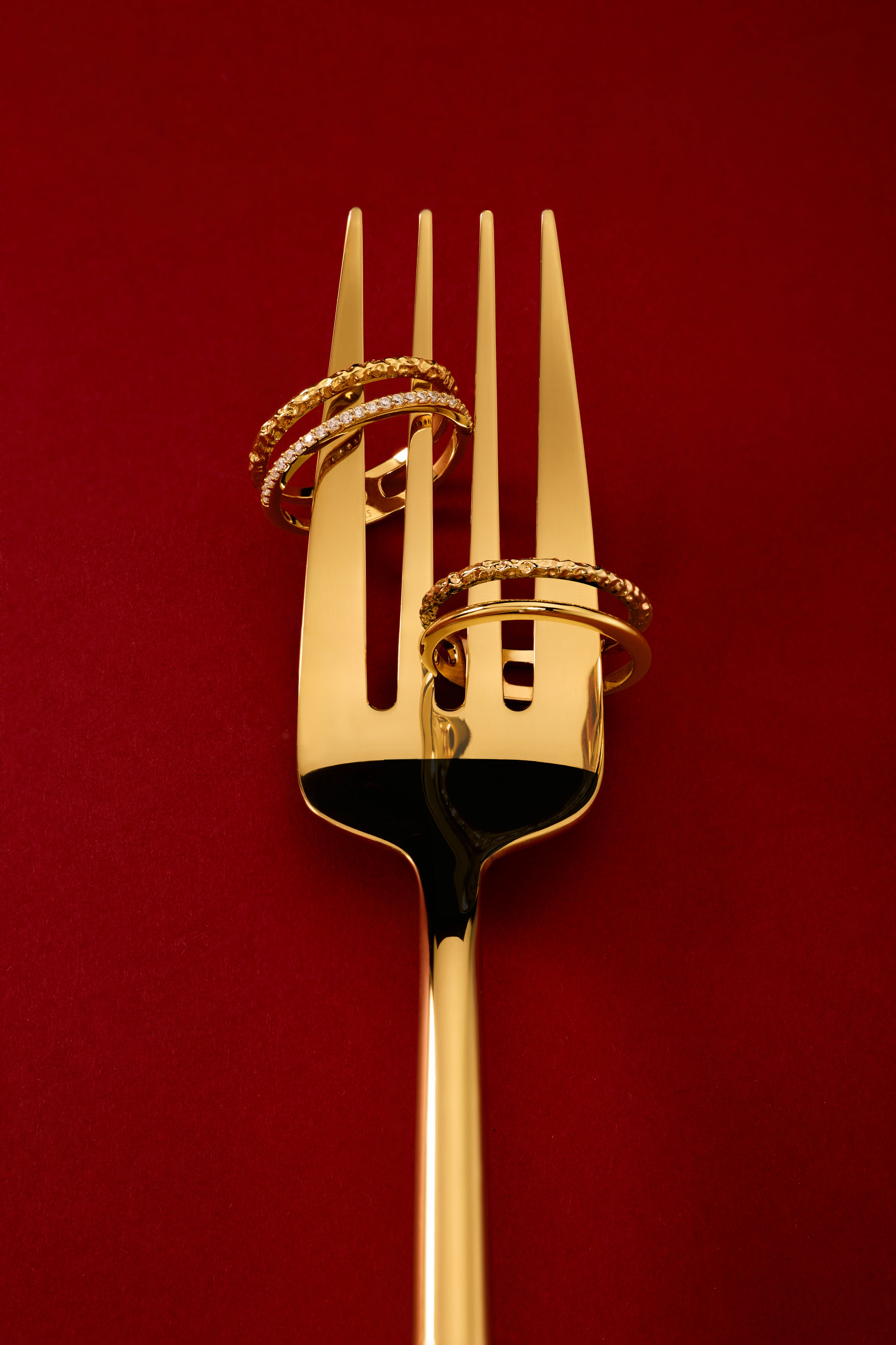 The Molten Crystal Double Band Ring in Gold, featuring two 18k gold plated bands, is elegantly displayed on a golden fork against a deep red background.
