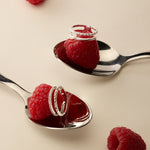Two silver spoons each hold a raspberry topped with the Molten Crystal Double Band Ring in Silver, while two more raspberries rest on the light beige surface nearby.