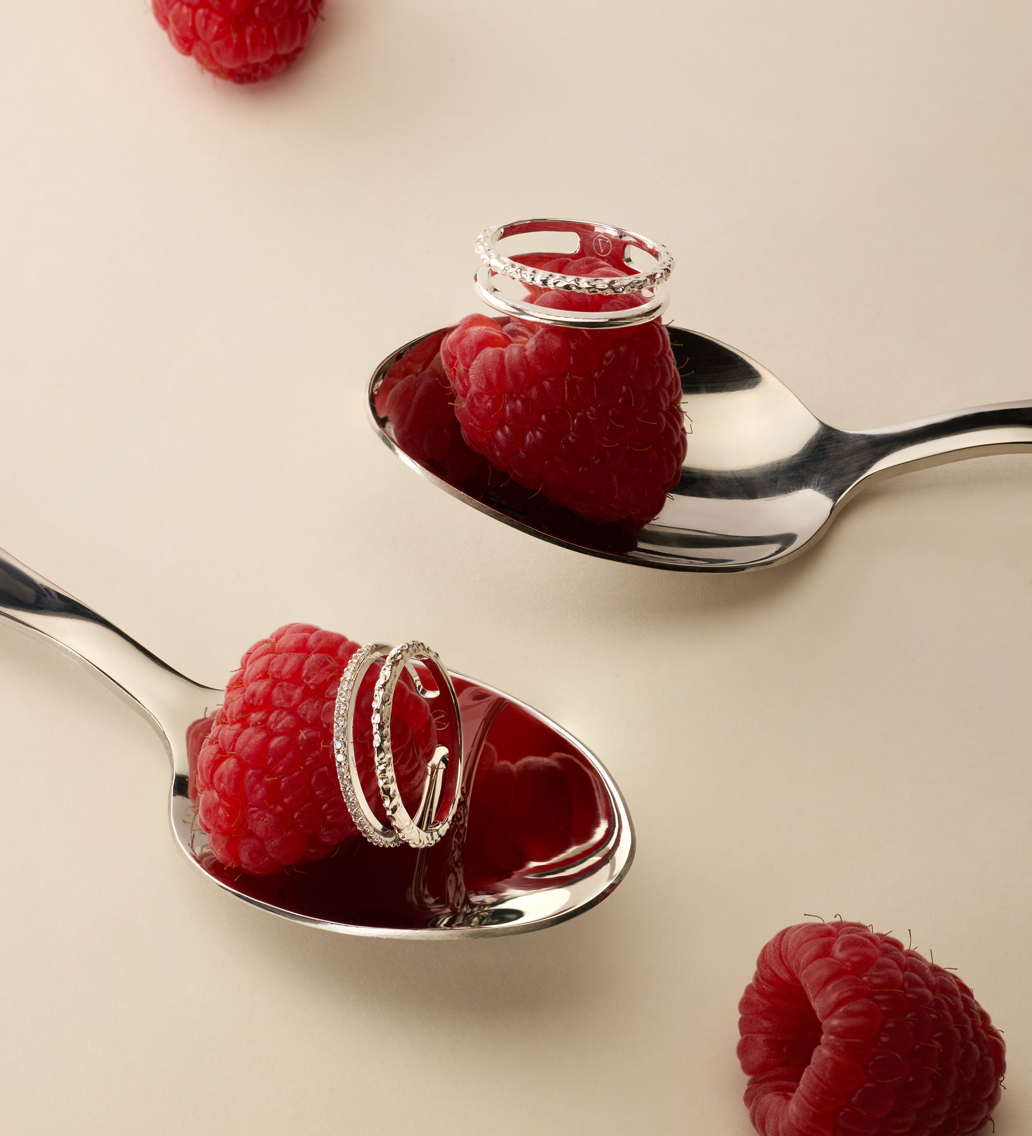 Two silver spoons each hold a raspberry topped with the Molten Crystal Double Band Ring in Silver, while two more raspberries rest on the light beige surface nearby.