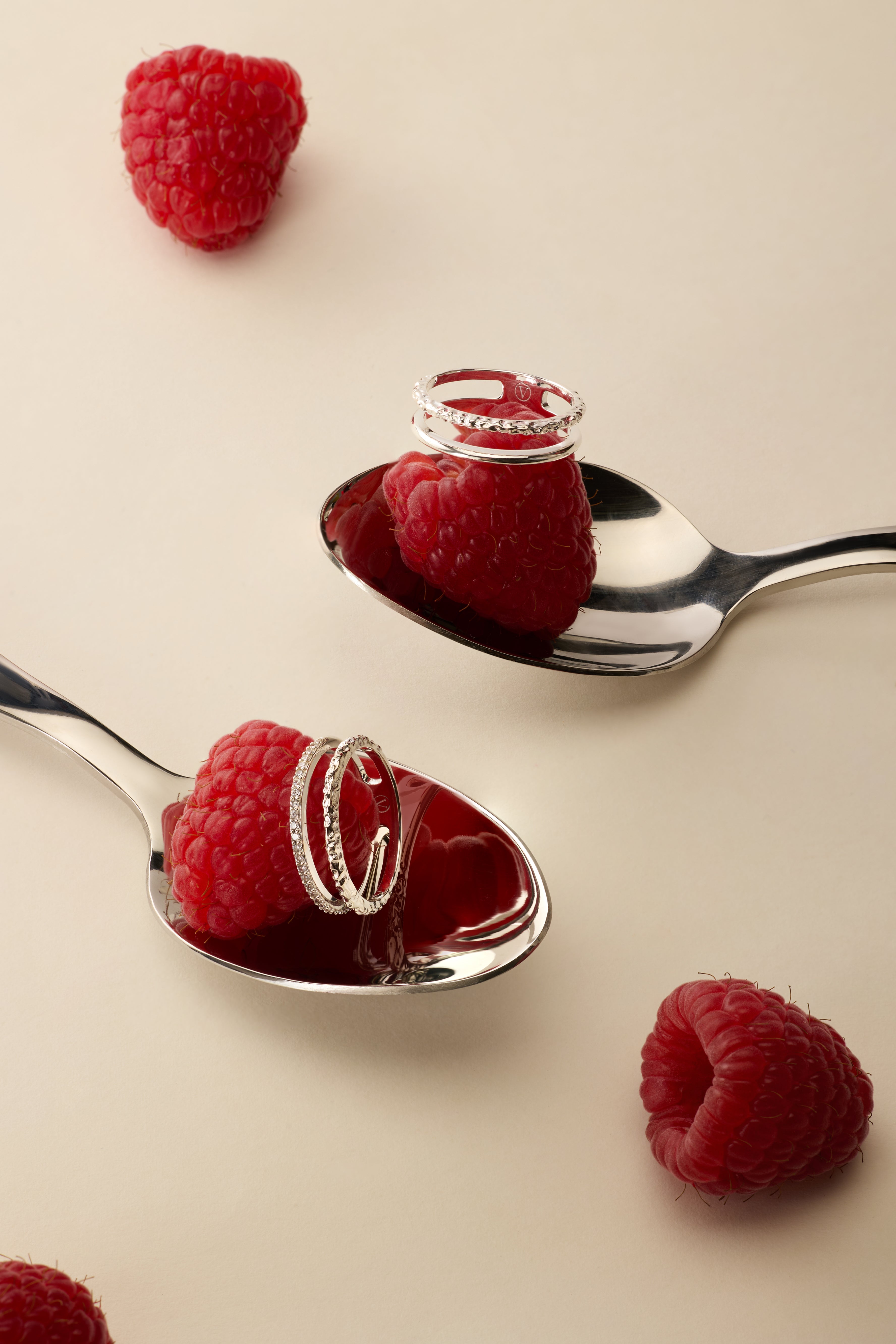 Two silver spoons each hold a raspberry topped with the Molten Crystal Double Band Ring in Silver, while two more raspberries rest on the light beige surface nearby.