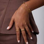 Wearing a brown outfit, a person rests their hand on their hip, showcasing 18k gold-plated bracelets, a Molten Crystal Double Band Ring in Gold with cubic zirconia, and neatly manicured light pink nails.
