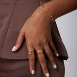 Wearing a brown outfit, a person rests their hand on their hip, showcasing 18k gold-plated bracelets, a Molten Crystal Double Band Ring in Gold with cubic zirconia, and neatly manicured light pink nails.