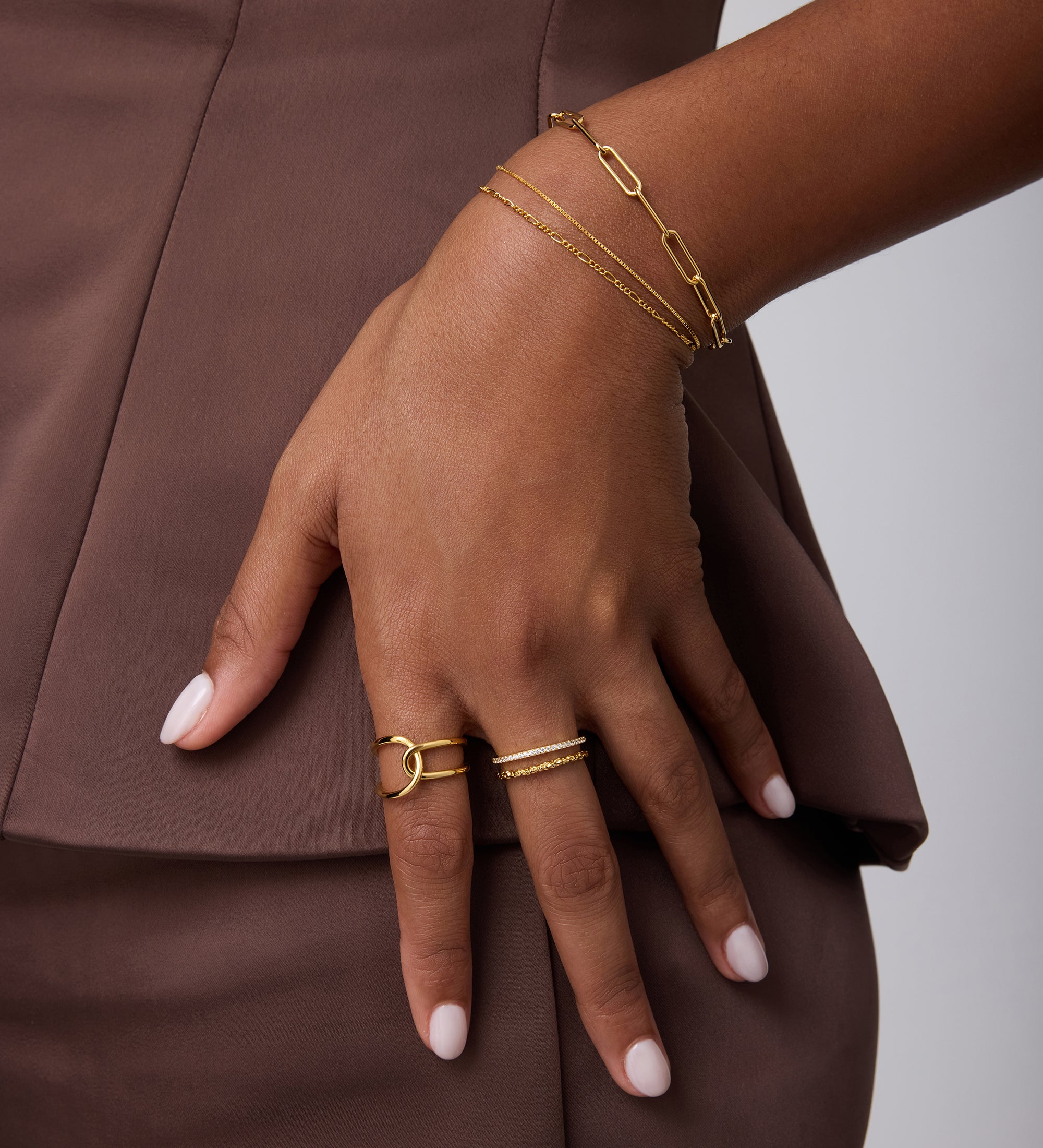 Wearing a brown outfit, a person rests their hand on their hip, showcasing 18k gold-plated bracelets, a Molten Crystal Double Band Ring in Gold with cubic zirconia, and neatly manicured light pink nails.