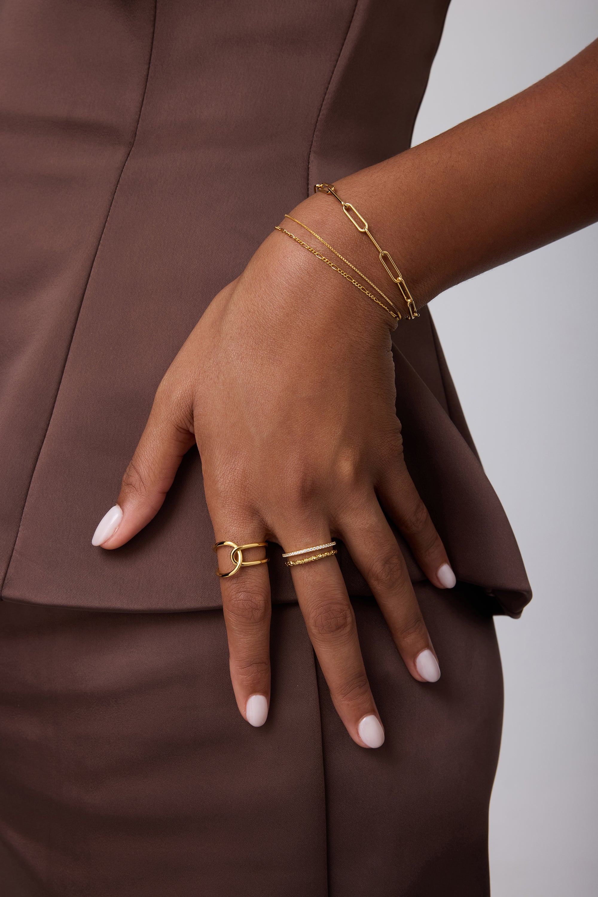 Wearing a brown outfit, a person rests their hand on their hip, showcasing 18k gold-plated bracelets, a Molten Crystal Double Band Ring in Gold with cubic zirconia, and neatly manicured light pink nails.