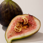 A halved fig with pink flesh holds two Nadine Pavé Twisted Stud Earrings in Gold, while a whole fig stands in the background, both illuminated by soft, natural light.