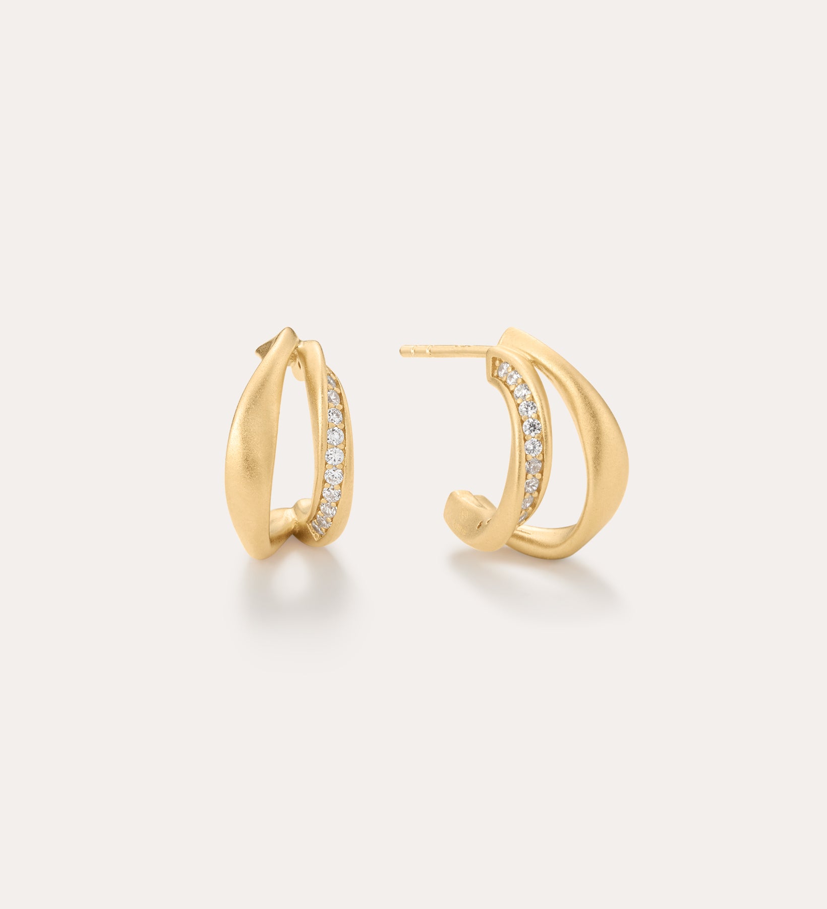 Nadine Pavé Twisted Stud Earrings in Gold feature a modern, sculptural design with a row of sparkling gemstones set along the inner edge and are displayed against a light neutral background.