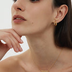 A woman with straight dark hair poses thoughtfully, resting her chin on her hand. She wears Nadine Pavé Twisted Stud Earrings in Gold and a delicate gold bar necklace, with minimal makeup against a plain, light background.