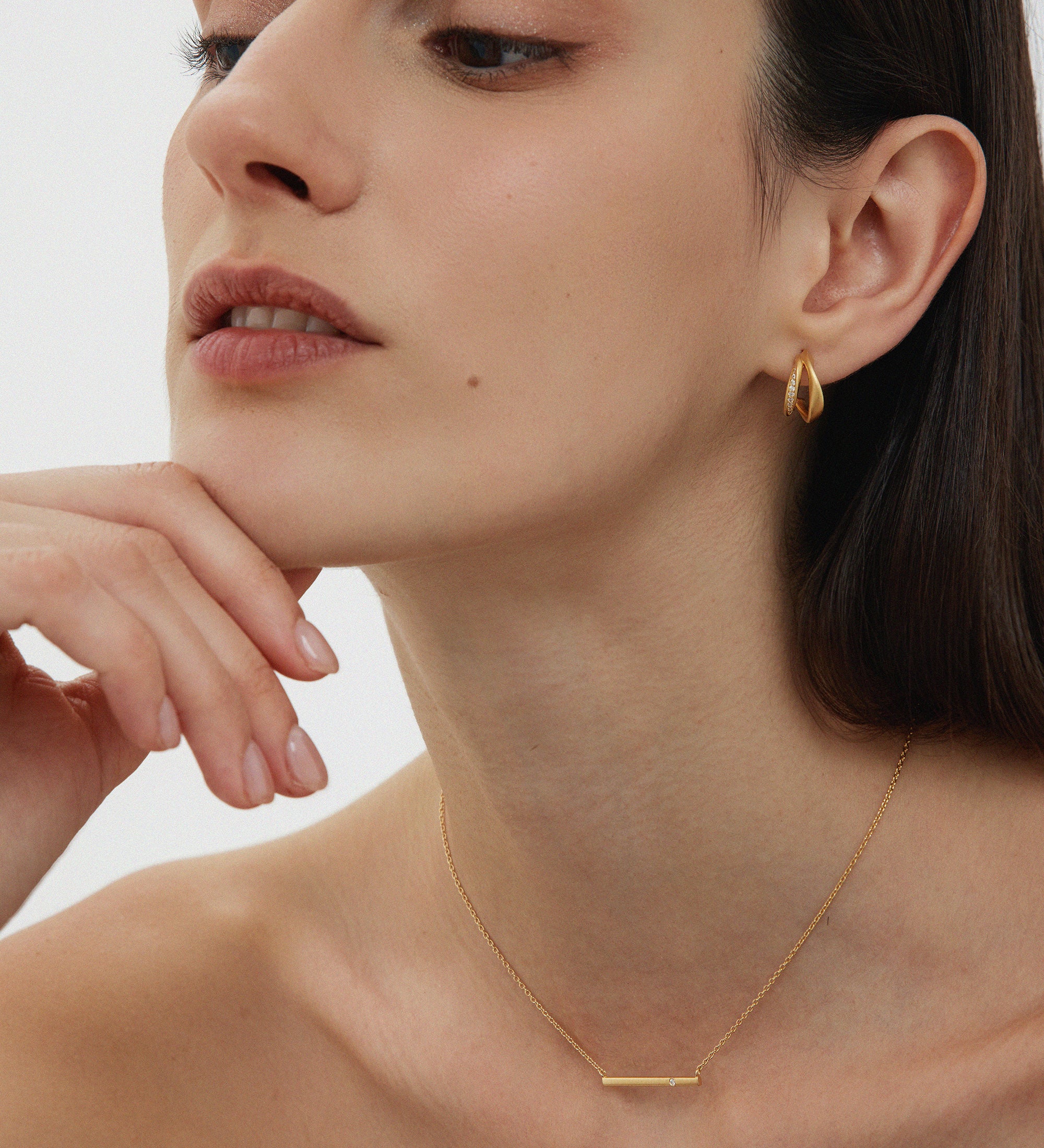 A woman with straight dark hair poses thoughtfully, resting her chin on her hand. She wears Nadine Pavé Twisted Stud Earrings in Gold and a delicate gold bar necklace, with minimal makeup against a plain, light background.