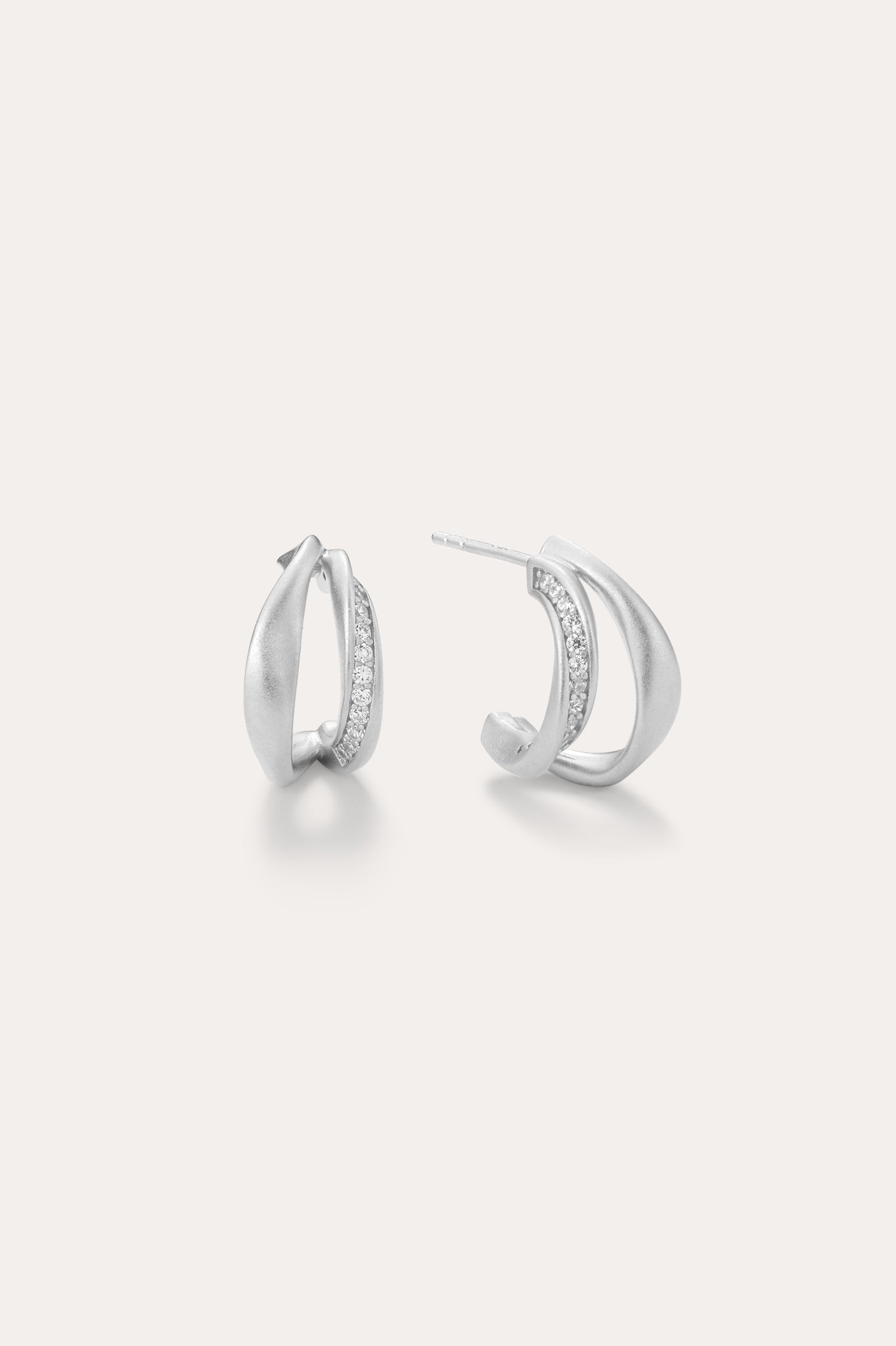 Nadine Pavé Twisted Stud Earrings in Silver feature lightweight, asymmetrical silver hoops with a row of sparkling stones along the inner curve, displayed on a plain white background.