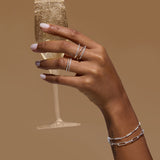 A hand with light pink nail polish wears the Nola Pavé Triple Band Ring in Silver and delicate bracelets while holding a glass of sparkling champagne against a plain brown background.