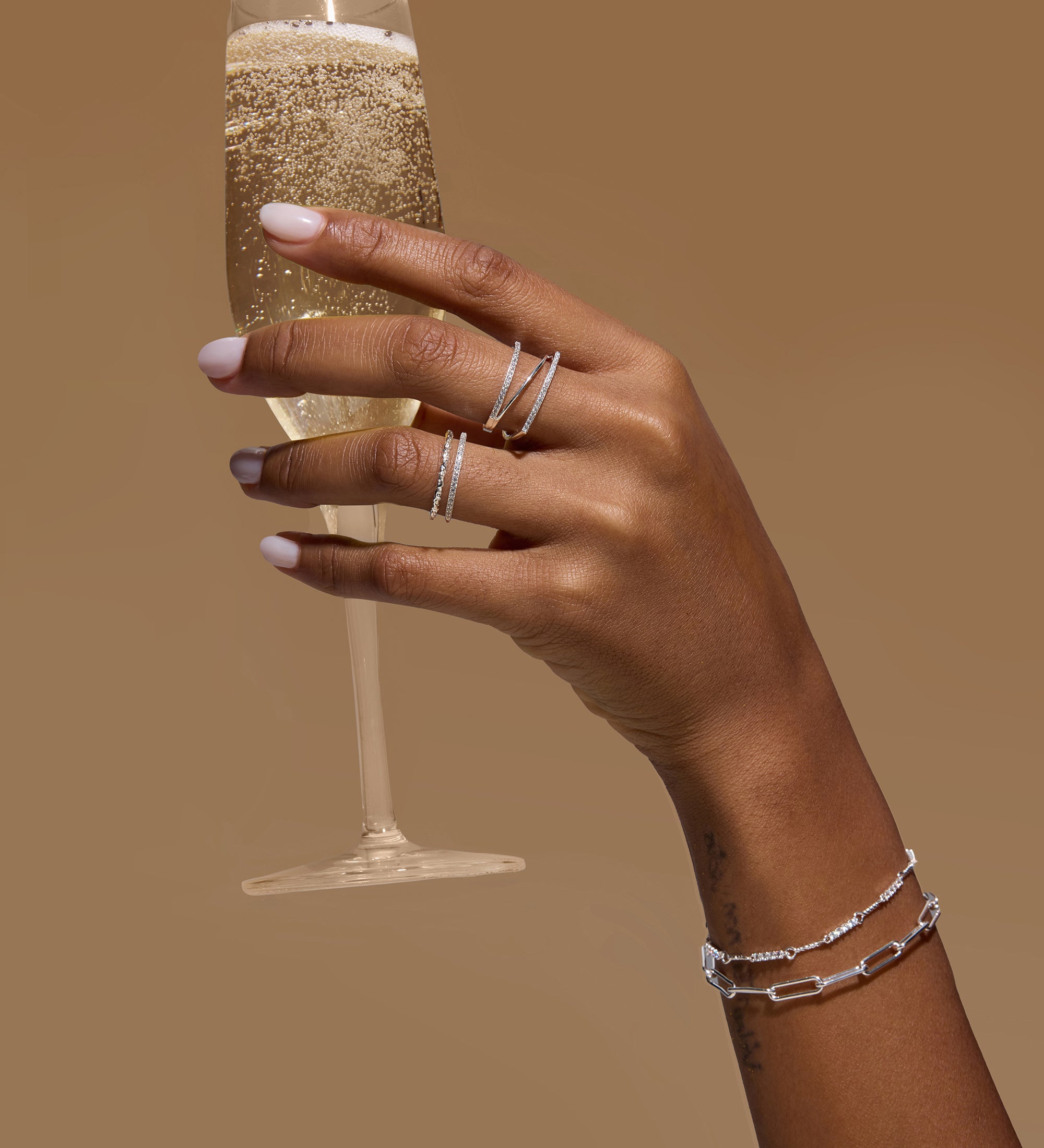 A hand with light pink nail polish wears the Nola Pavé Triple Band Ring in Silver and delicate bracelets while holding a glass of sparkling champagne against a plain brown background.