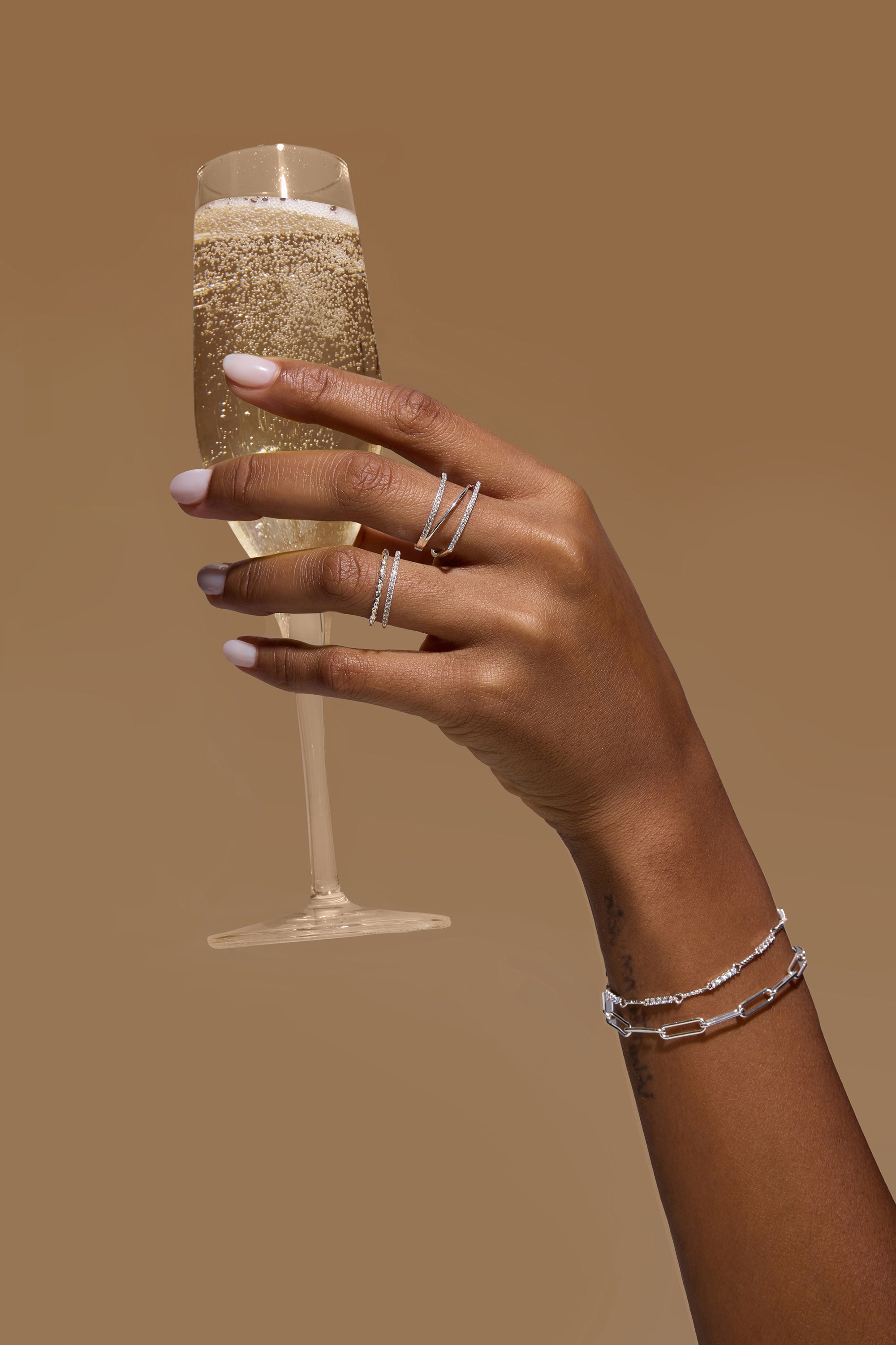 A hand with light pink nail polish wears the Nola Pavé Triple Band Ring in Silver and delicate bracelets while holding a glass of sparkling champagne against a plain brown background.