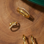 The Noor Pavé Triple Band Ring in Gold, along with three other gold jewelry pieces including twisted hoop earrings, is displayed on a smooth wooden surface beside a green circular object.