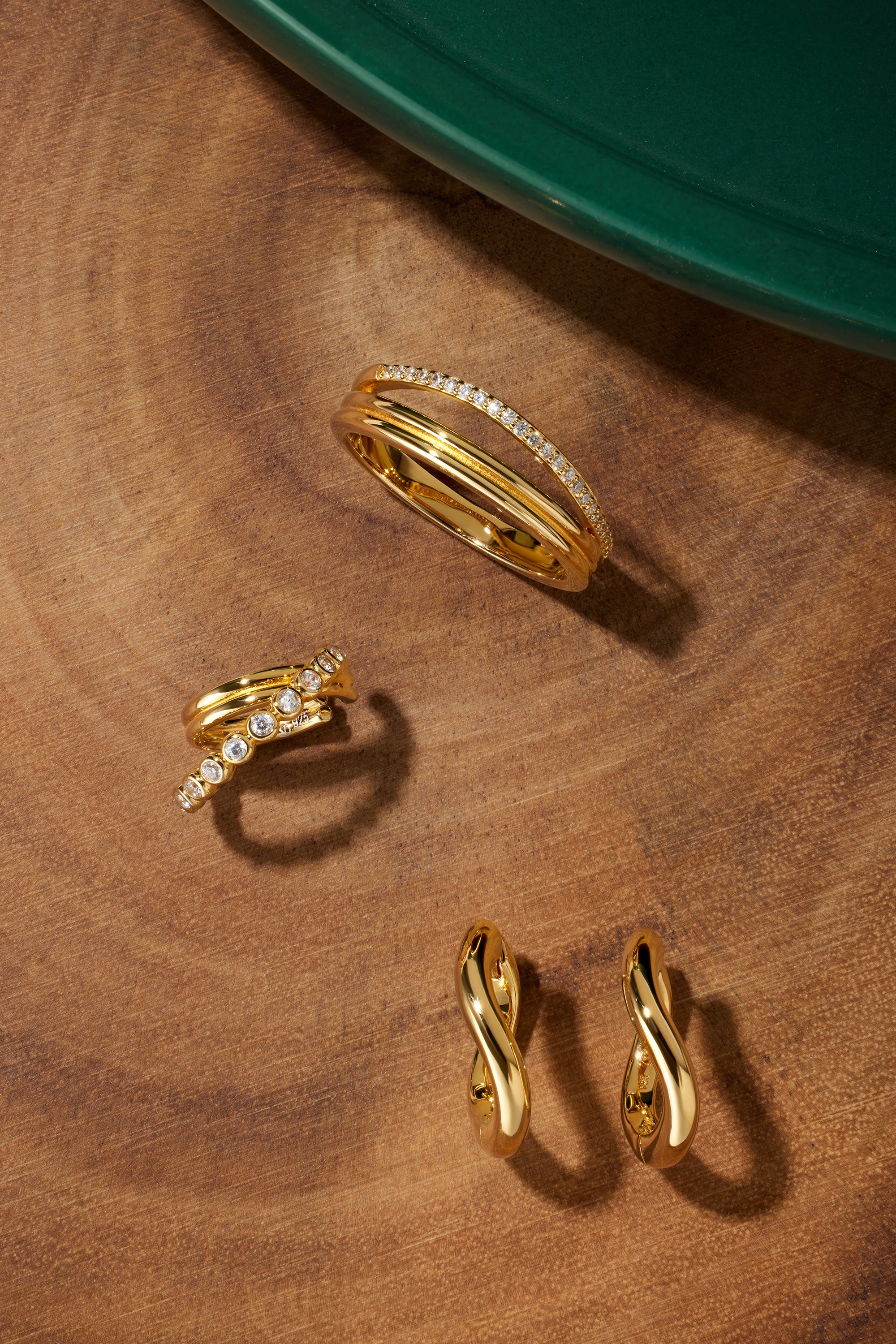 The Noor Pavé Triple Band Ring in Gold, along with three other gold jewelry pieces including twisted hoop earrings, is displayed on a smooth wooden surface beside a green circular object.