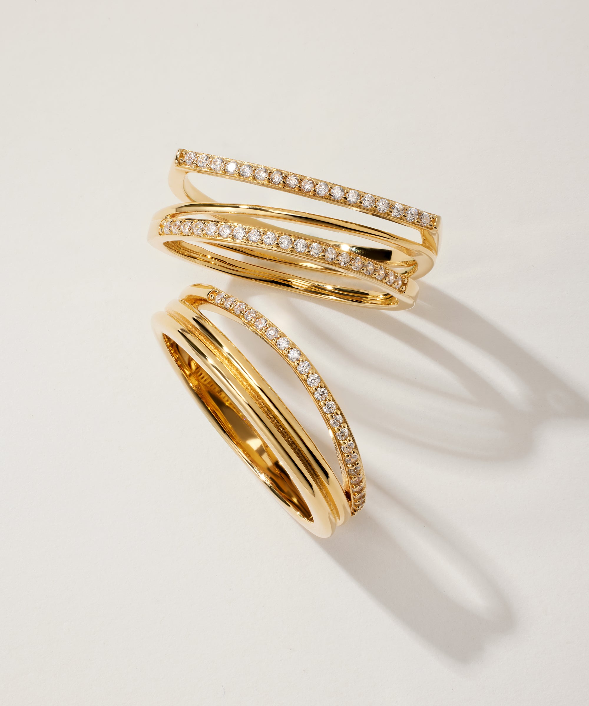 The Noor Pavé Triple Band Ring in Gold features overlapping pavé bands, each set with a row of sparkling diamonds, displayed on a light background with soft shadows.