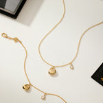 The Nora Double Pendant Bracelet in Gold, featuring sculptural heart and teardrop charms, is displayed on a light surface with scattered books and artistically designed cards.