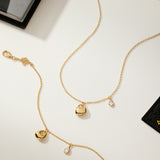 The Nora Double Pendant Bracelet in Gold, featuring sculptural heart and teardrop charms, is displayed on a light surface with scattered books and artistically designed cards.