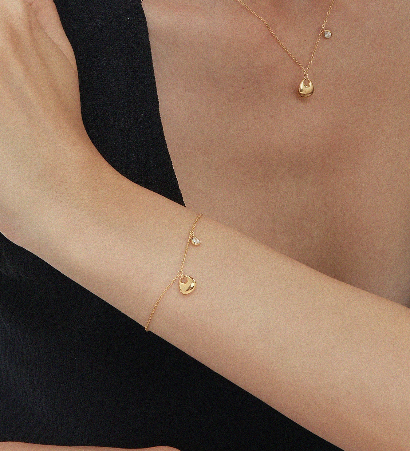 A person in a sleeveless black top rests their arm on a table, showcasing the Nora Double Pendant Bracelet in Gold alongside other gold jewelry, with their hand touching their upper arm against a plain white background.