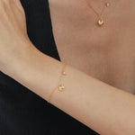 A person in a sleeveless black top rests their arm on a table, showcasing the Nora Double Pendant Bracelet in Gold alongside other gold jewelry, with their hand touching their upper arm against a plain white background.
