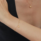 A person in a sleeveless black top rests their arm on a table, showcasing the Nora Double Pendant Bracelet in Gold alongside other gold jewelry, with their hand touching their upper arm against a plain white background.