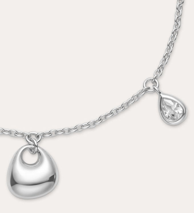 The Nora Double Pendant Bracelet in Silver features an adjustable clasp, a small heart-shaped charm, and a tiny cubic zirconia teardrop charm dangling side by side.