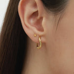 Close-up of a womans ear adorned with two earrings: a small flower stud and the Nori Twisted Hoop Earrings in Gold. The neutral background highlights the elegant shine of these 18k gold plated hoops.