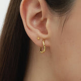 Close-up of a womans ear adorned with two earrings: a small flower stud and the Nori Twisted Hoop Earrings in Gold. The neutral background highlights the elegant shine of these 18k gold plated hoops.