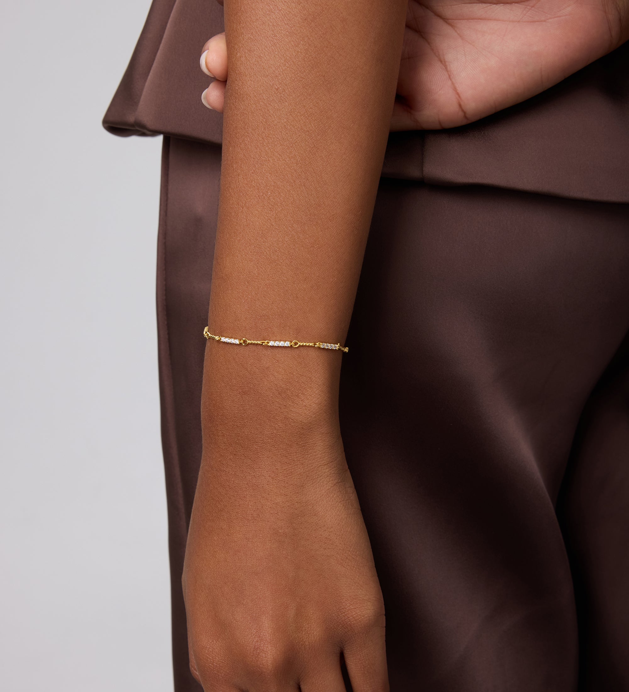 A person in a brown satin outfit rests their left arm by their side, showing off the Pia Crystal Link Bracelet in Gold on their wrist against a plain, neutral background.