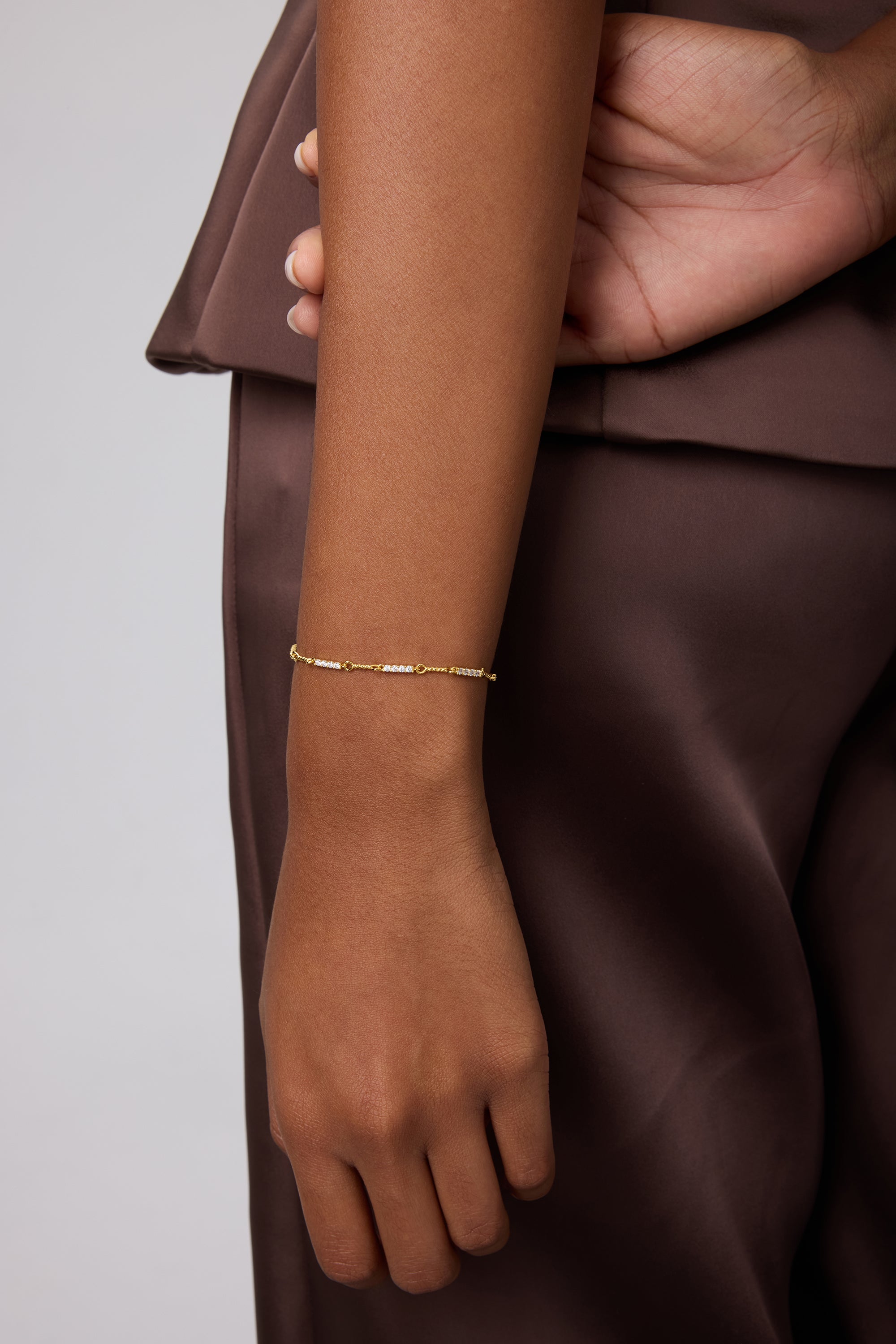 A person in a brown satin outfit rests their left arm by their side, showing off the Pia Crystal Link Bracelet in Gold on their wrist against a plain, neutral background.
