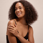 A woman with curly hair, dressed in a black tank top and gold jewelry, smiles while displaying the Reine Rainbow Crystal Ring in Gold on her hand against a plain light background.