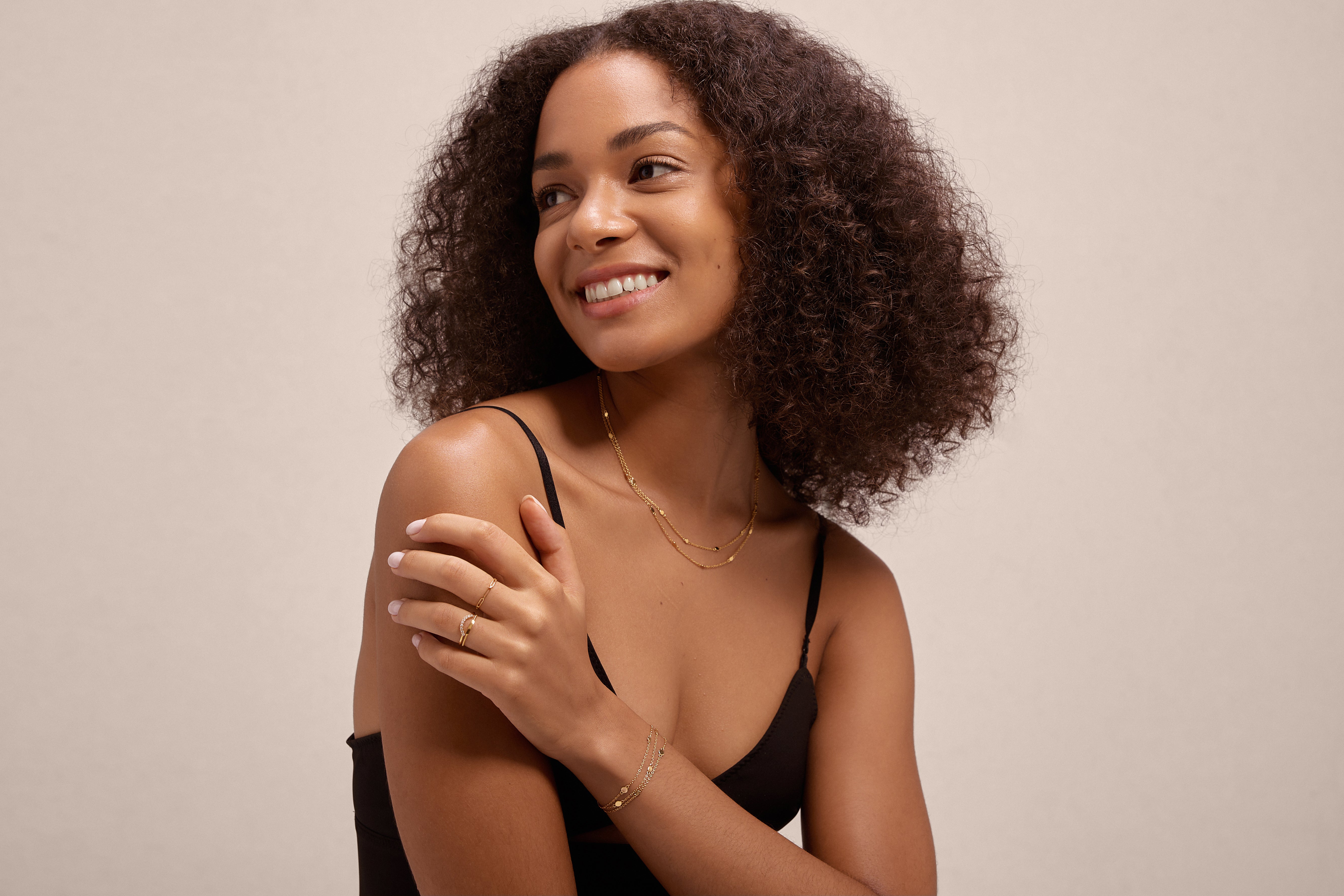 A woman with curly hair, dressed in a black tank top and gold jewelry, smiles while displaying the Reine Rainbow Crystal Ring in Gold on her hand against a plain light background.