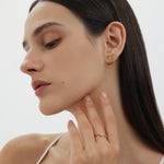 A woman with straight dark hair and natural makeup looks down, wearing Reneé Triangle Linear Hoop Stud Earrings in Gold and a gold ring, her hand gently touching her neck against a plain white background.