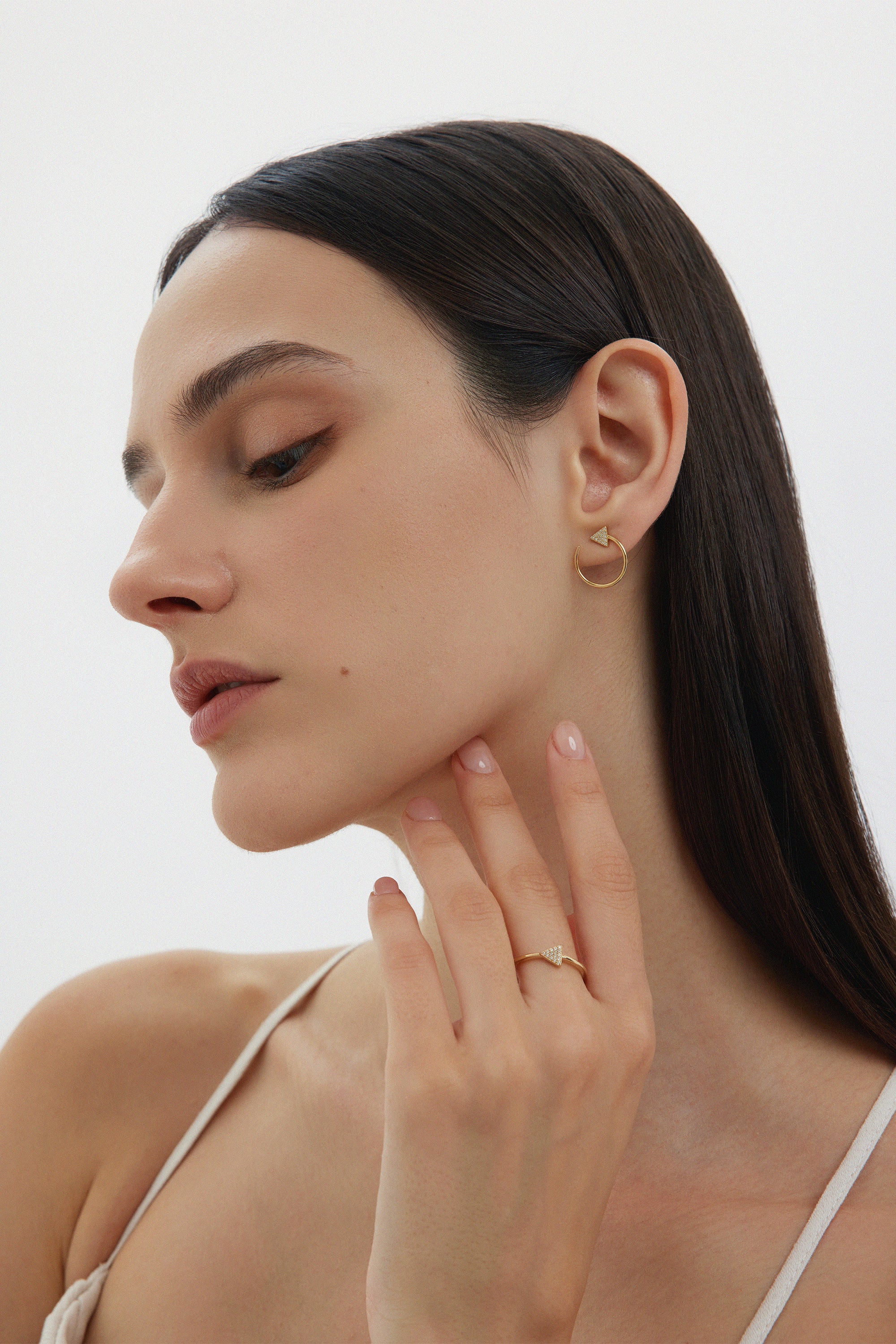 A woman with straight dark hair and natural makeup looks down, wearing Reneé Triangle Linear Hoop Stud Earrings in Gold and a gold ring, her hand gently touching her neck against a plain white background.