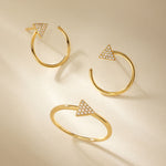 Three Reneé Triangle Linear Hoop Stud Earrings in Gold, each adorned with a diamond-studded triangle, are artfully arranged on a beige background with soft shadows.