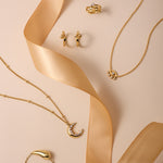 Gold Ribbon Twist Huggies are displayed on a beige surface with a satin ribbon, alongside gold necklaces featuring crescent moon pendants and delicate chains for an elegant, stylish look.