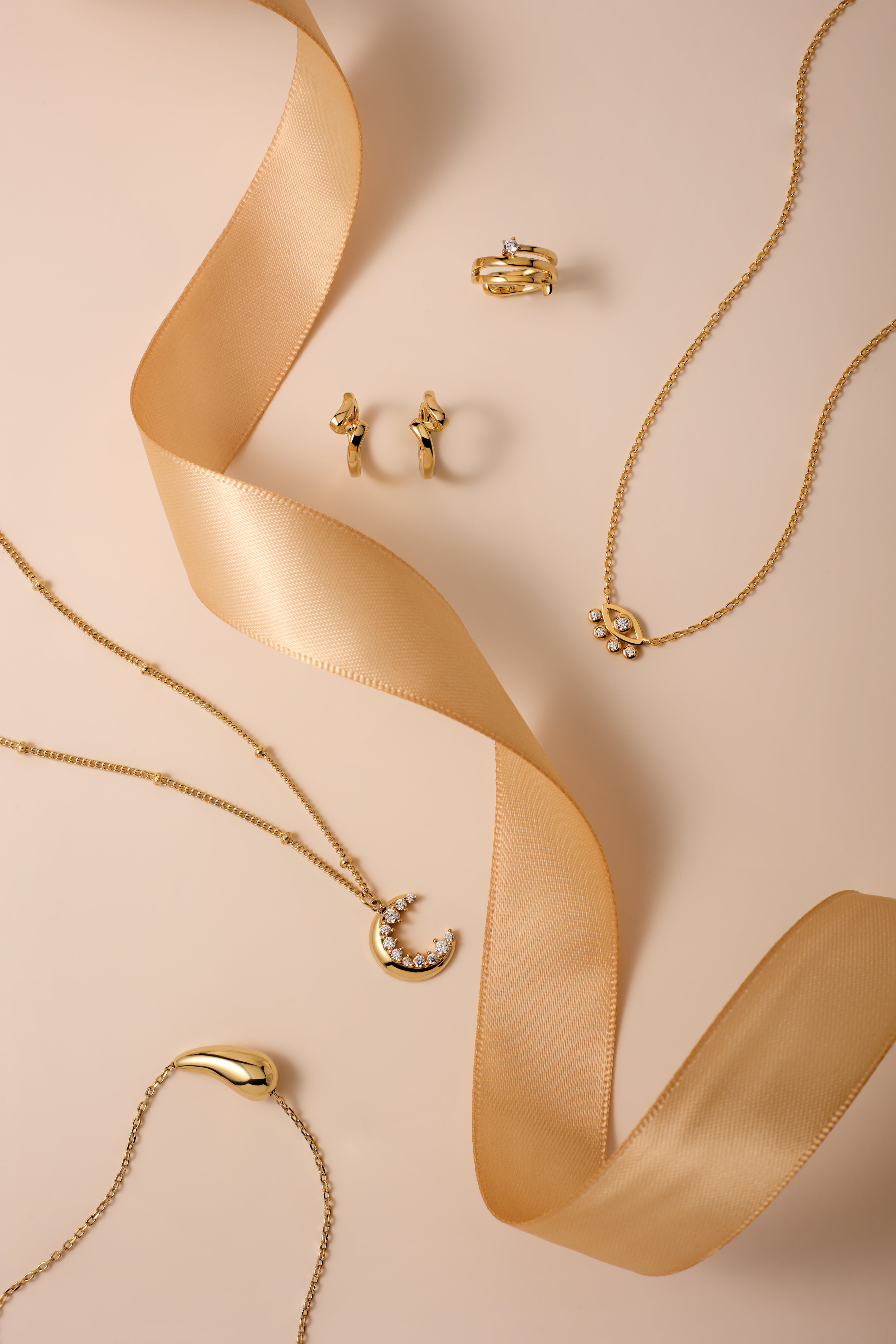 Gold Ribbon Twist Huggies are displayed on a beige surface with a satin ribbon, alongside gold necklaces featuring crescent moon pendants and delicate chains for an elegant, stylish look.