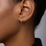 Close-up of a person wearing the Ribbon Twist Huggies in Silver—small sterling silver hoop earrings featuring a delicate twisted ribbon design. The individual has medium-brown skin, dark hair pulled back, and wears a black top against a light background.