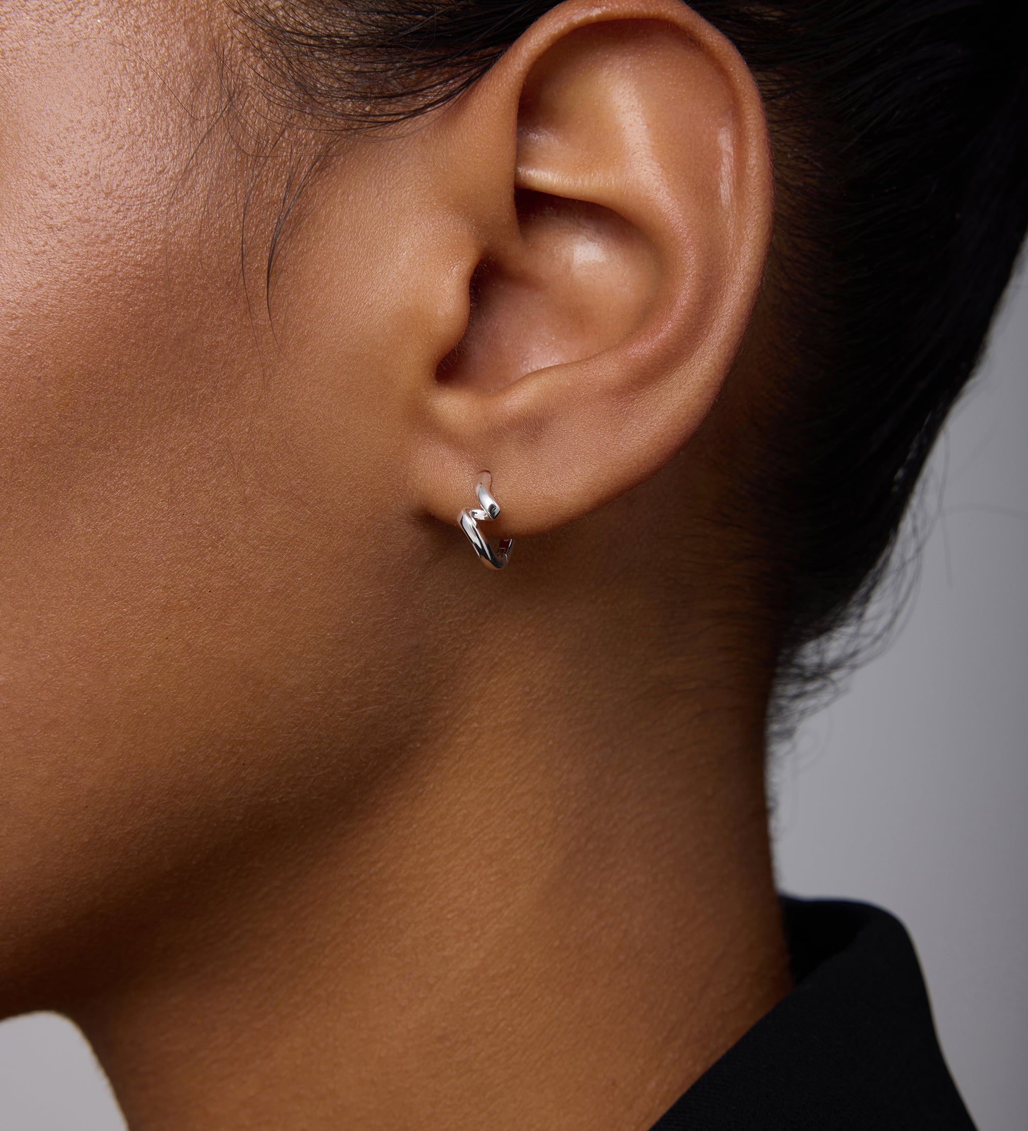 Close-up of a person wearing the Ribbon Twist Huggies in Silver—small sterling silver hoop earrings featuring a delicate twisted ribbon design. The individual has medium-brown skin, dark hair pulled back, and wears a black top against a light background.