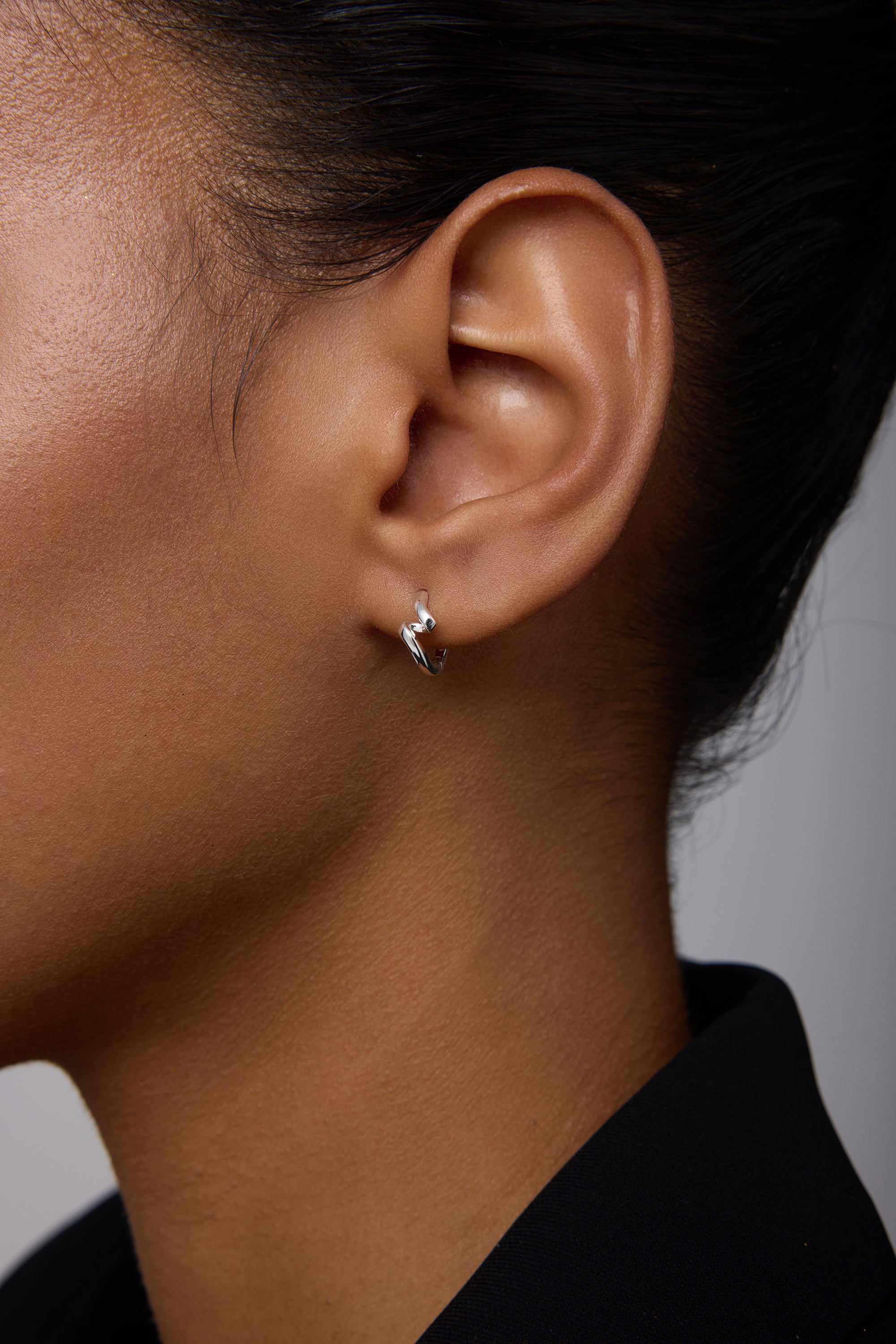 Close-up of a person wearing the Ribbon Twist Huggies in Silver—small sterling silver hoop earrings featuring a delicate twisted ribbon design. The individual has medium-brown skin, dark hair pulled back, and wears a black top against a light background.