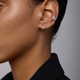 Close-up of a person’s ear wearing Ribbon Twist Huggies in Silver on the lobe and another silver huggie on the upper ear. The person has smooth skin and is dressed in a black blazer.